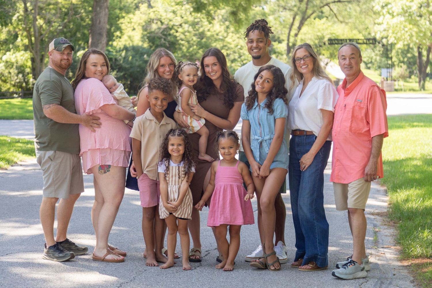 A diverse family group poses joyfully outdoors at Kinnick Feller Riverside Park in Adel, Iowa, surrounded by lush trees.