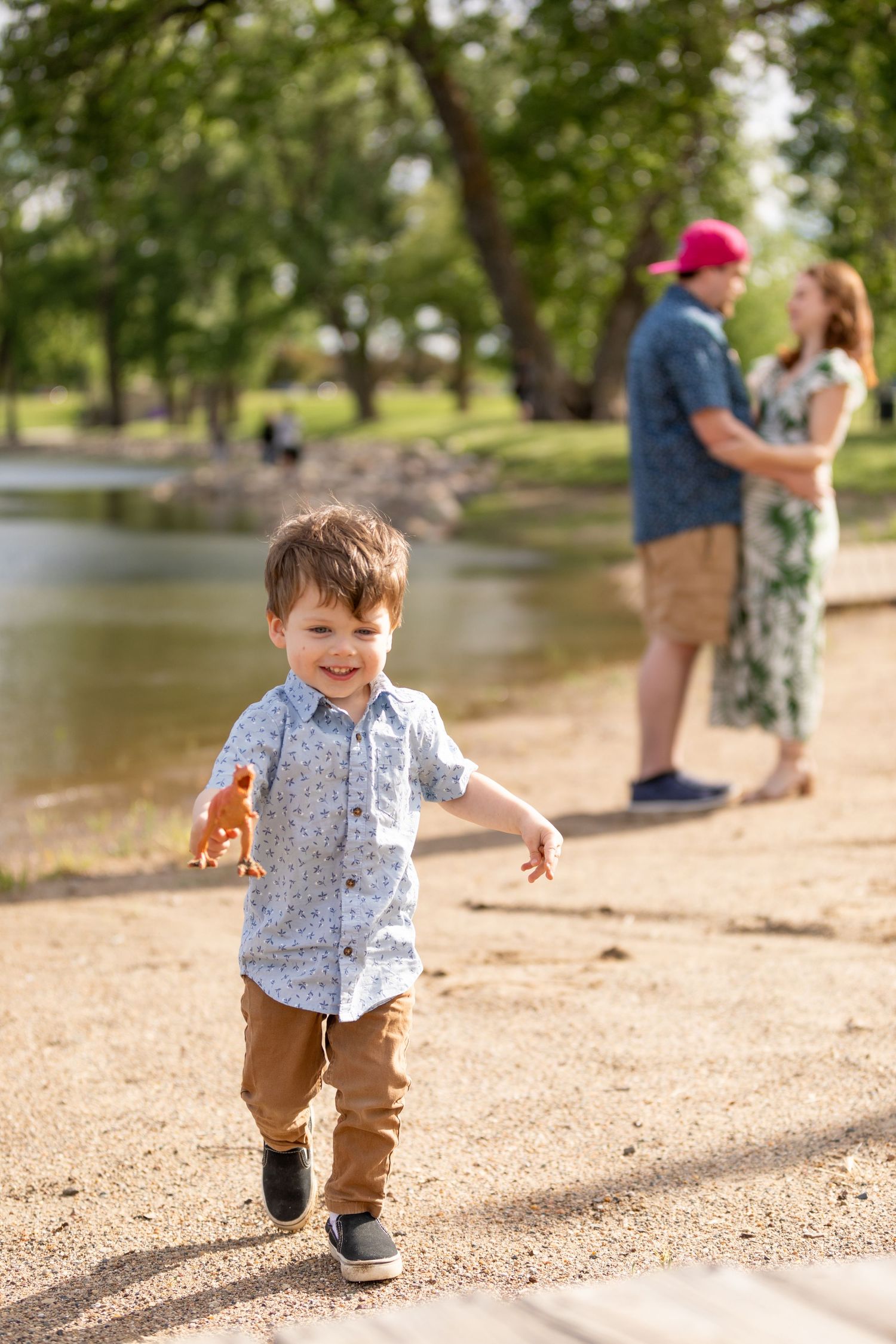 A joyful toddler runs in the foreground on a sunny day. Behind him, a couple shares a kiss at Gray's Lake Park in Des Moines, Iowa.