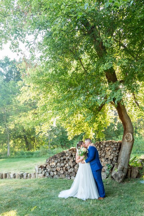 A bride and groom share a kiss under a large tree in a sunlit garden. The bride wears a white gown, and the groom is in a blue suit. Logs ar