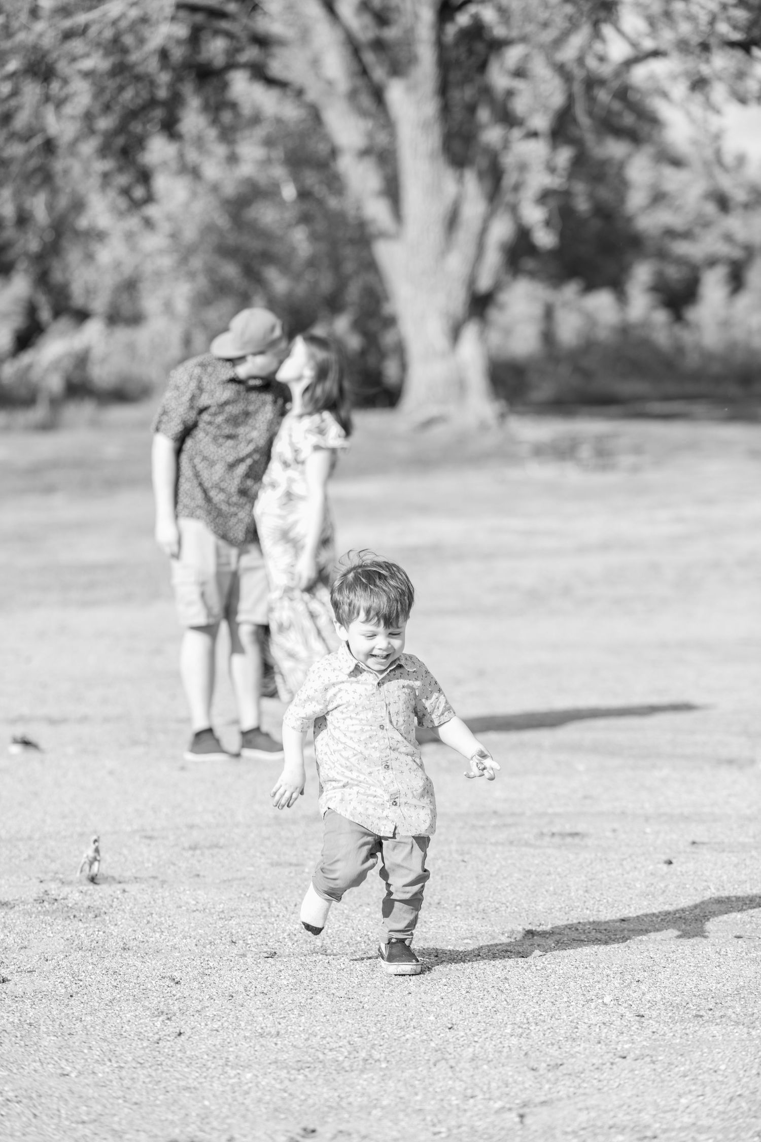 A joyful toddler runs in the foreground on a sunny day. Behind him, a couple shares a kiss at Gray's Lake Park in Des Moines, Iowa.