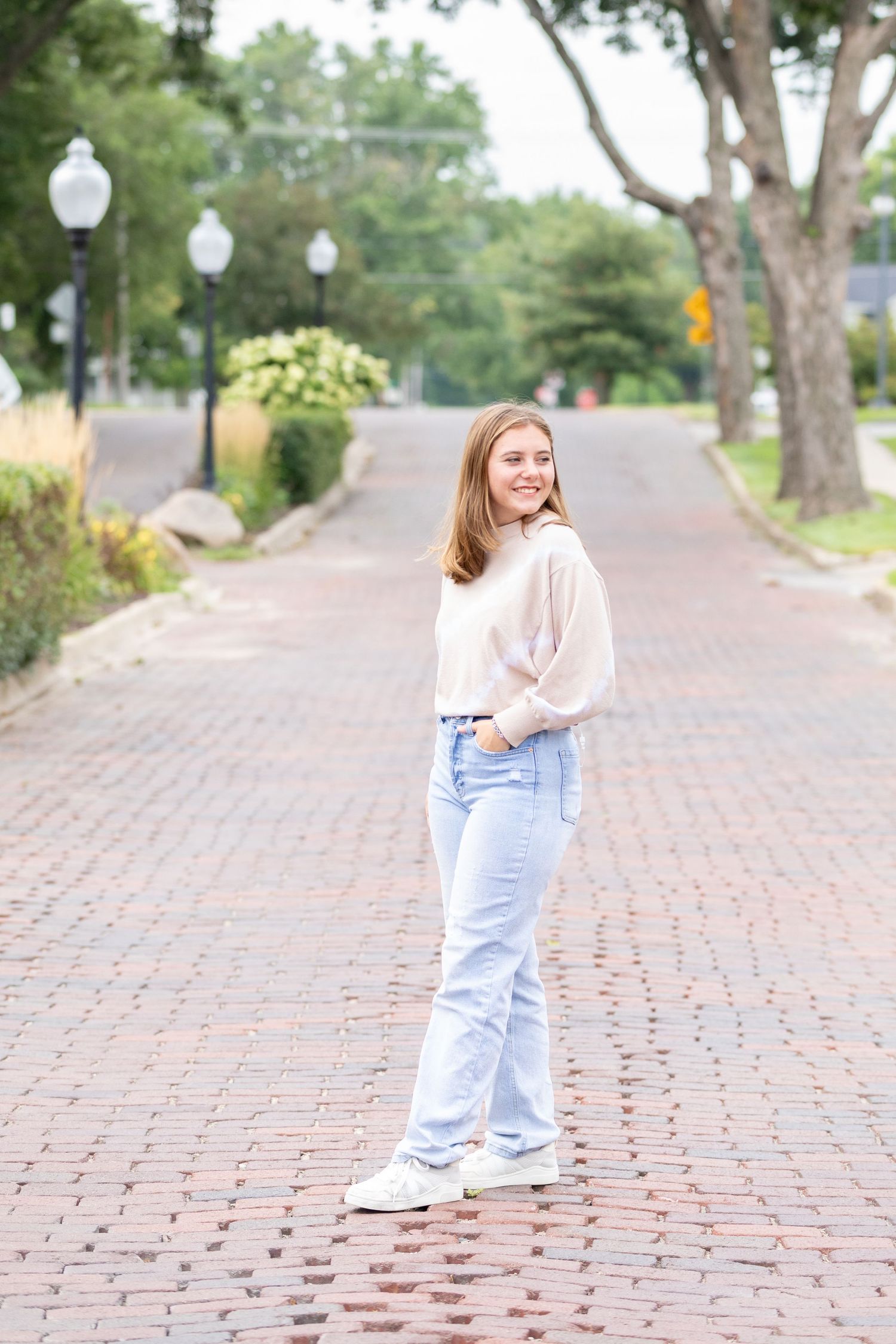 A high school senior in light jeans and a cream sweater stands on a brick road in Adel, Iowa under tree-lined street with soft lighting.