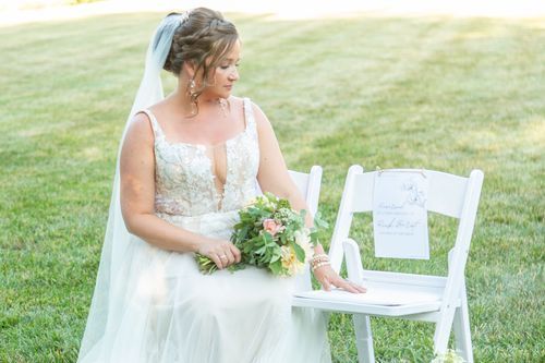 A bride in a white gown with a veil sits holding a bouquet, touching an empty chair with a sign. The scene feels serene and reflective.