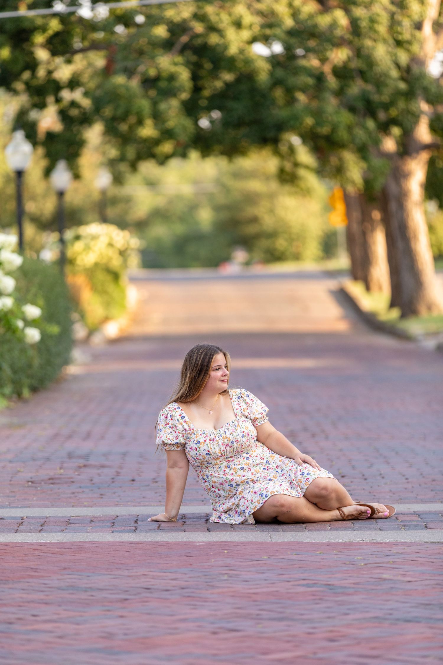 A high school senior in a floral dress sits on a brick road in Adel, Iowa under tree-lined street with soft, warm lighting.