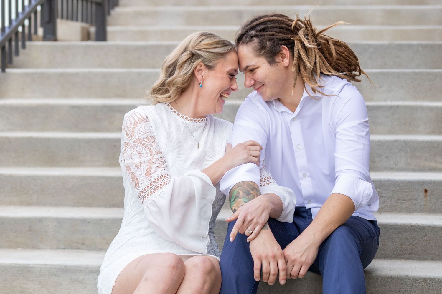 A couple sits on steps in Des Moines, IA smiling and leaning their heads together.