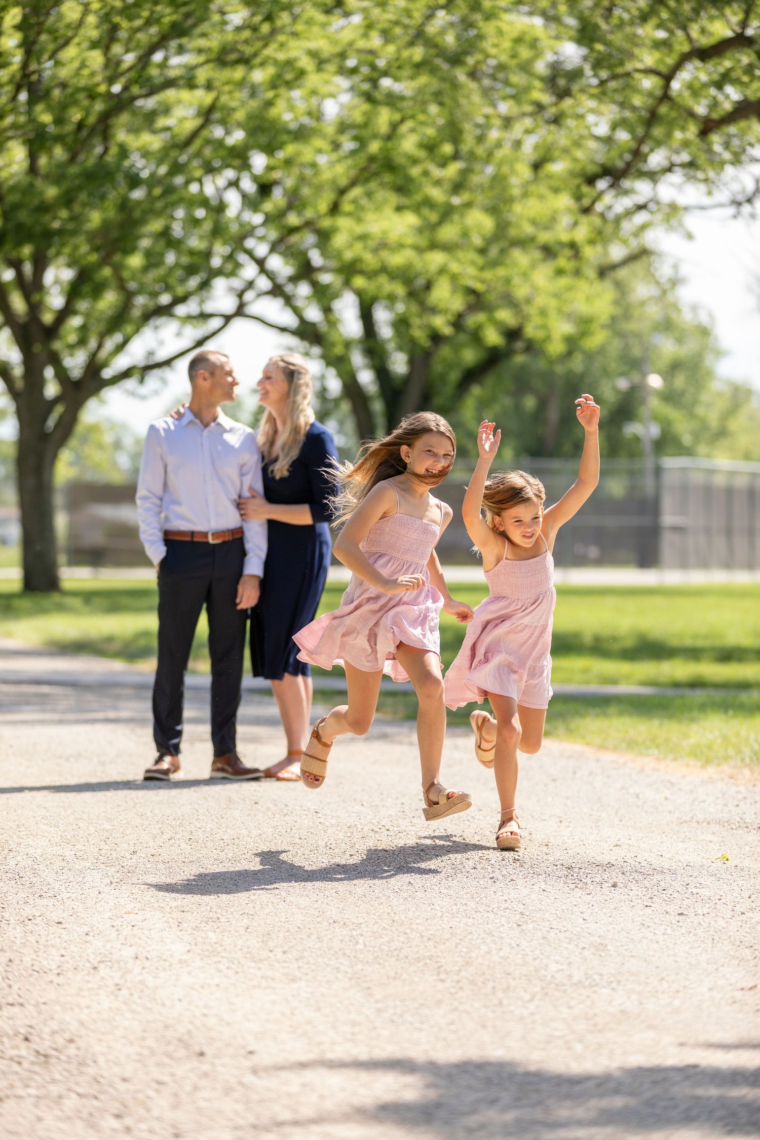 Two young girls laugh and run down a sunlit path at Kinnick Feller Riverside Park in Adel, Iowa with a couple smiling in the background.