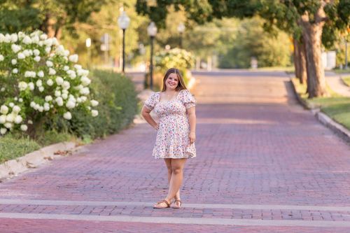 A high school senior in a floral dress stands confidently on a sunlit, brick-paved street lined with trees and blooming bushes in Adel, Iowa