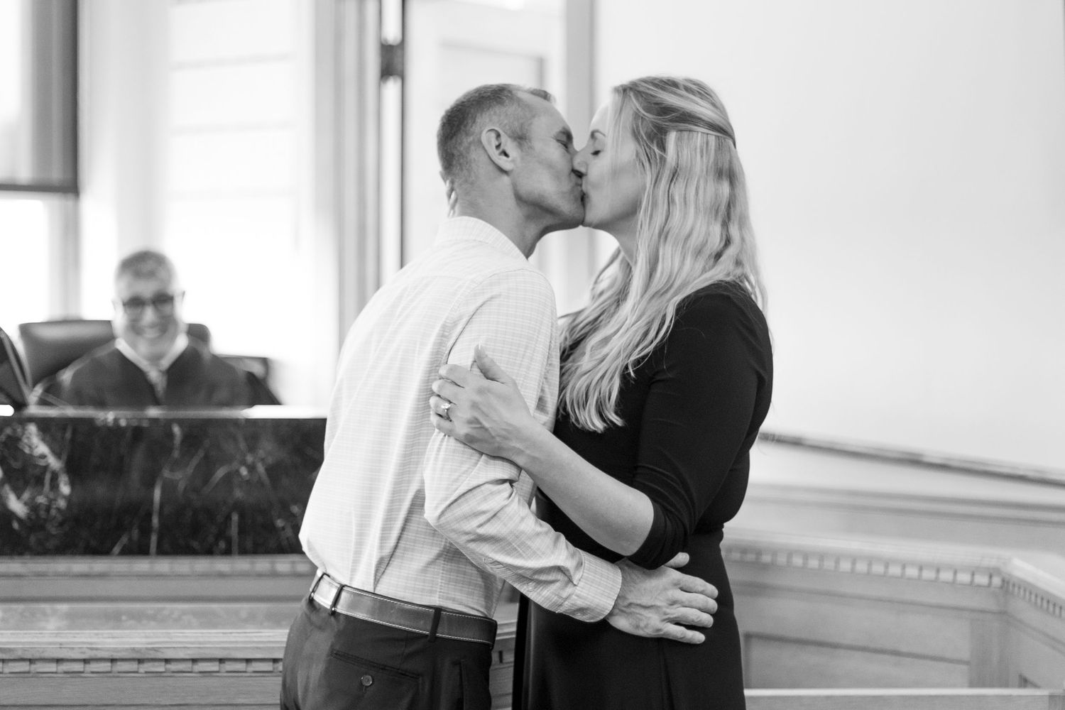 A couple shares their first kiss at the Dallas County Iowa Courthouse while a smiling officiant observes in the back.
