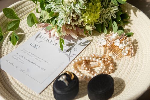 A wedding invitation with floral design lies on a woven mat, surrounded by pearl jewelry, a gold ring in a black box, and a bouquet.