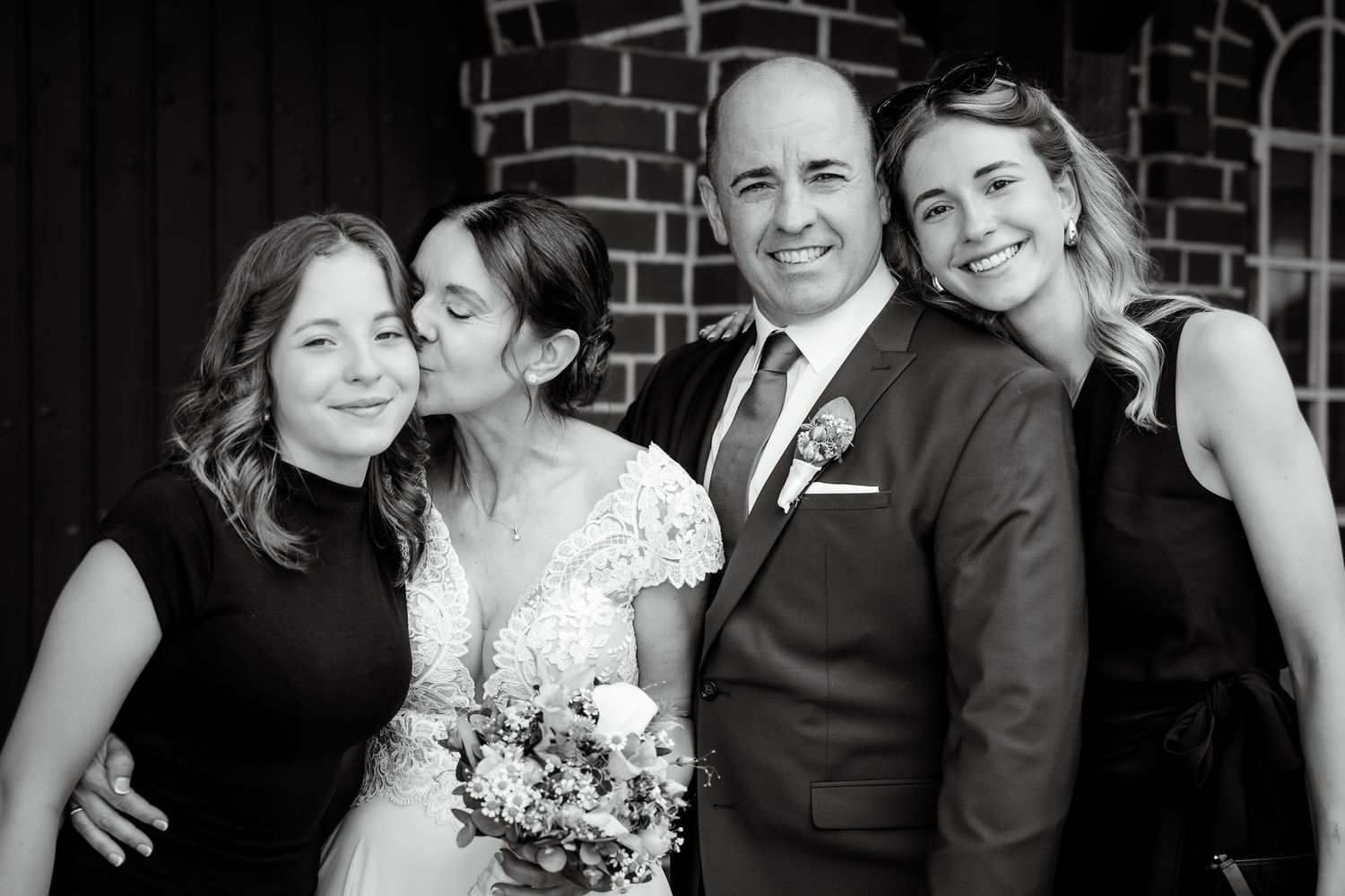 Black and white photo of a bride kissing the groom on the cheek while two women smile beside them.