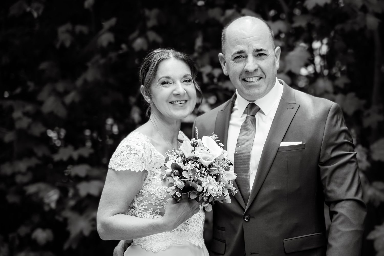 Black and white wedding photo of smiling bride holding bouquet and groom in suit posing outdoors.