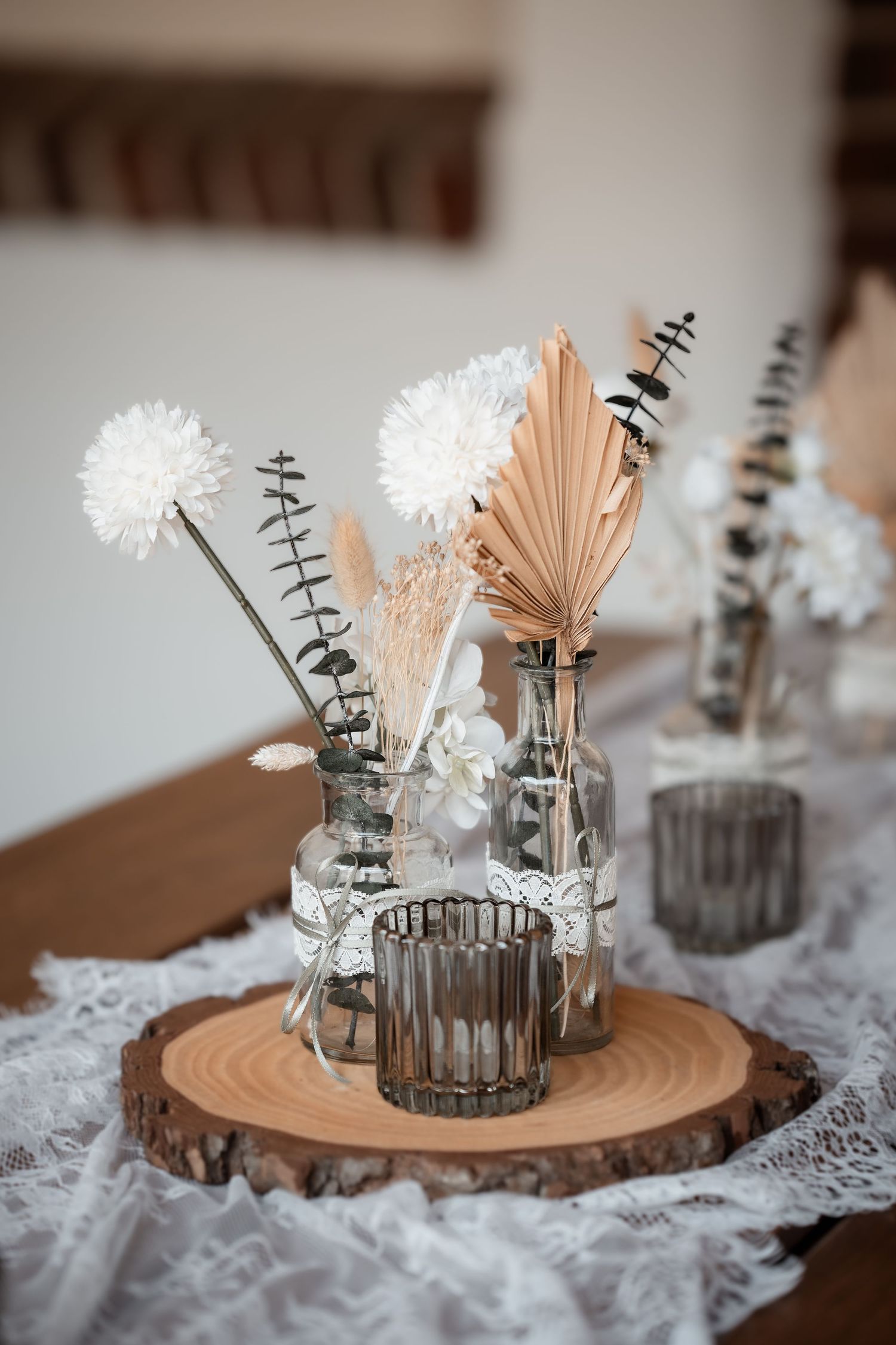 Elegant boho wedding table centerpiece with dried flowers in glass bottles on a wooden slice with lace runner.