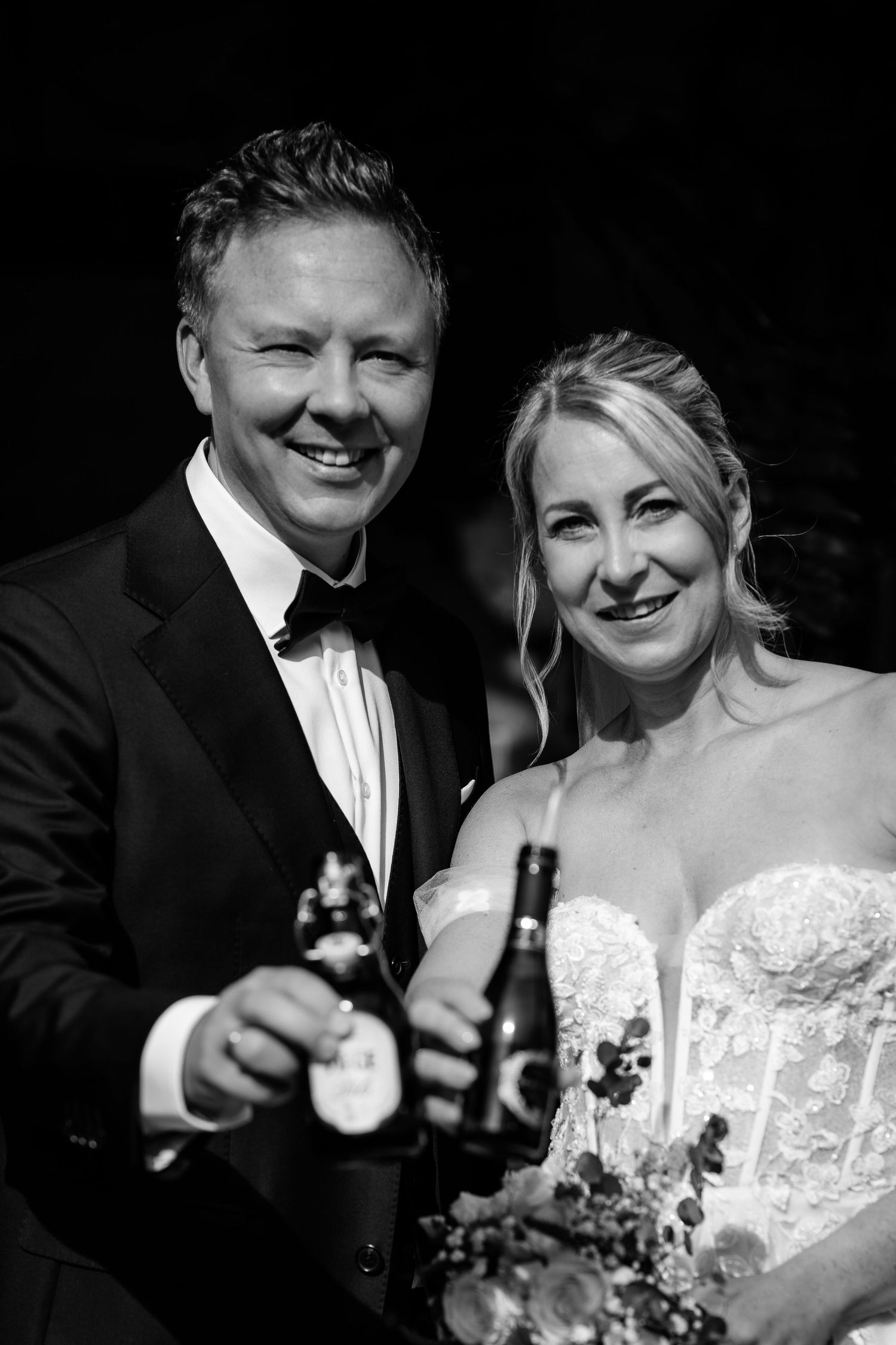Smiling bride and groom in tuxedo toasting with beer bottles at their wedding reception.