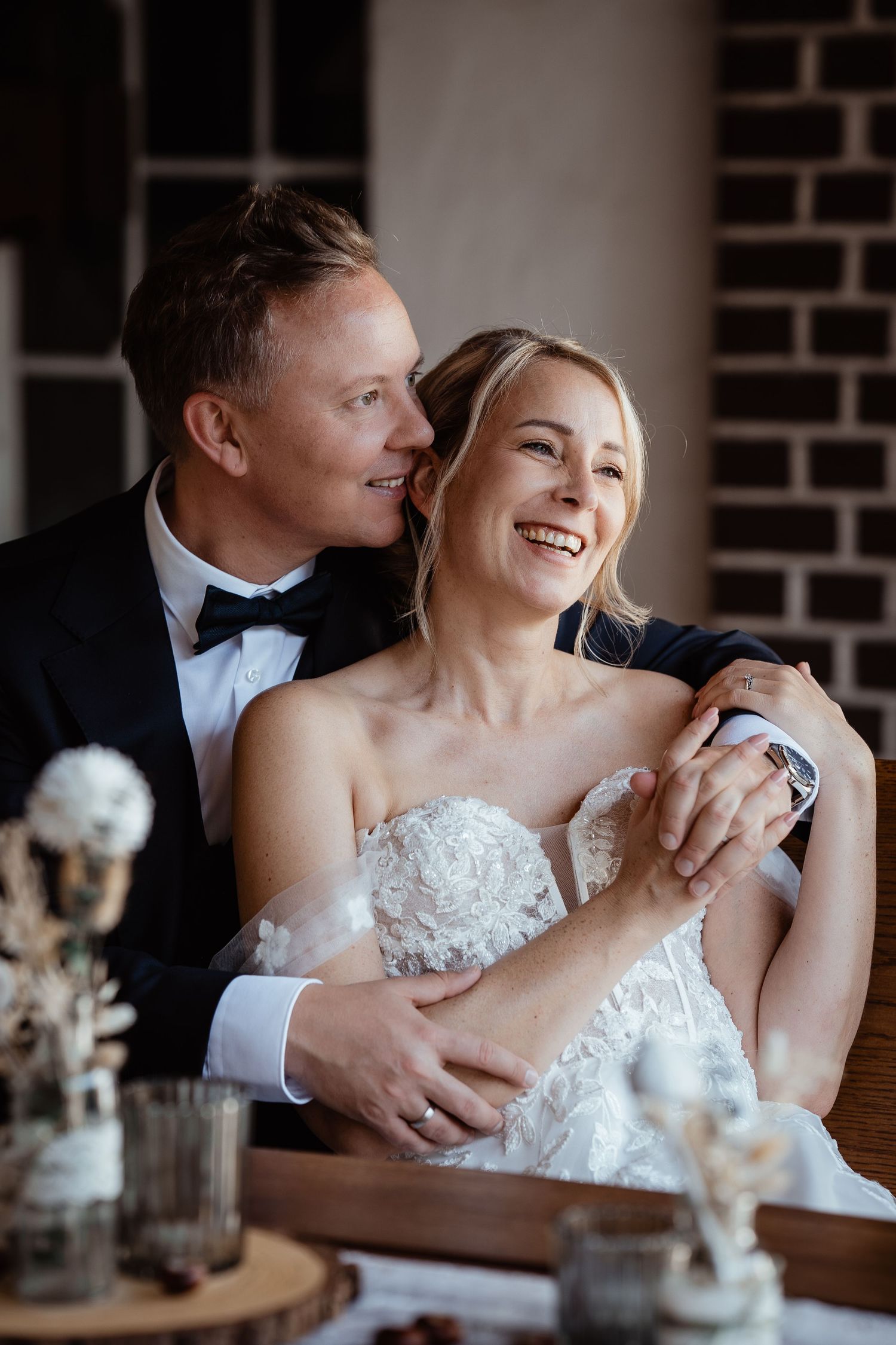 Smiling bride in lace gown embraced by groom in tuxedo seated at wedding reception table.