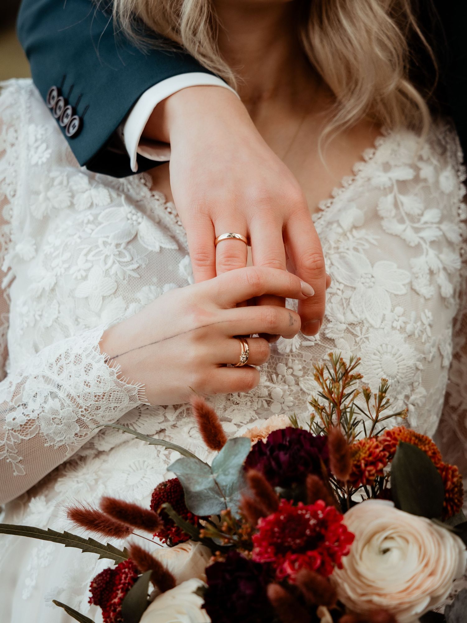 Close-up of bride and groom's hands showing wedding rings, with a red and white bridal bouquet.