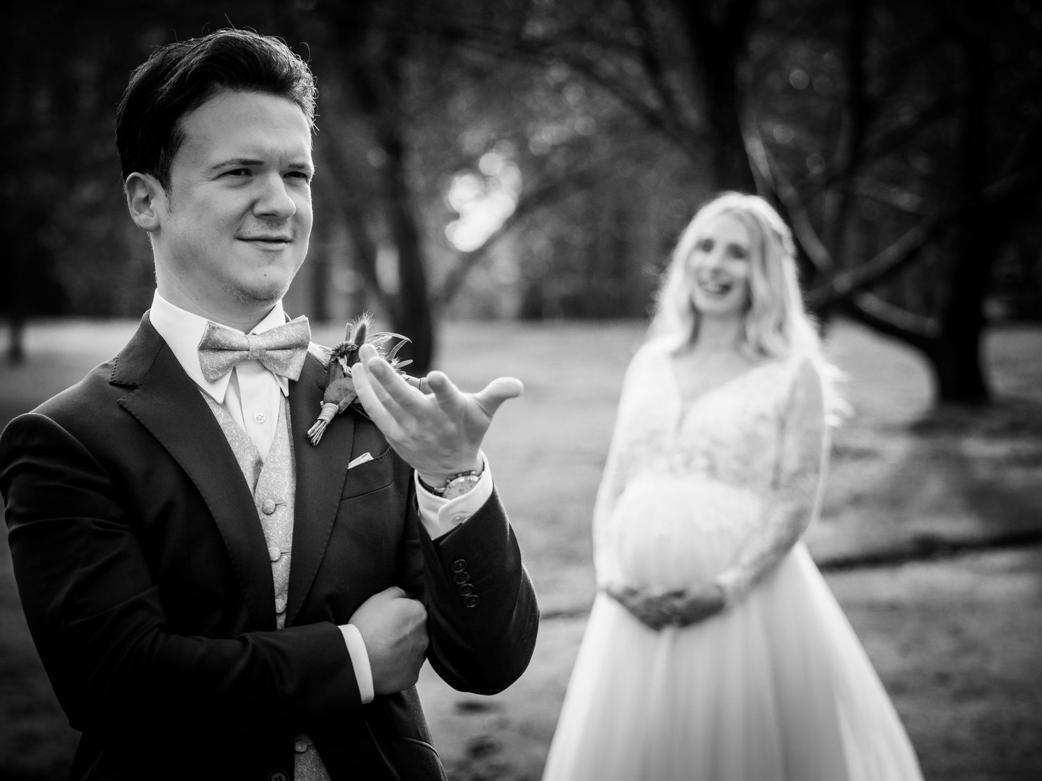 Black and white wedding photo of groom smiling with pregnant bride standing softly blurred in background.