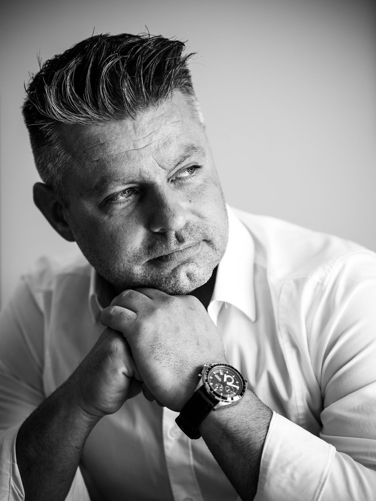 Black and white portrait of a contemplative person in a white shirt wearing a watch, resting chin on hands.