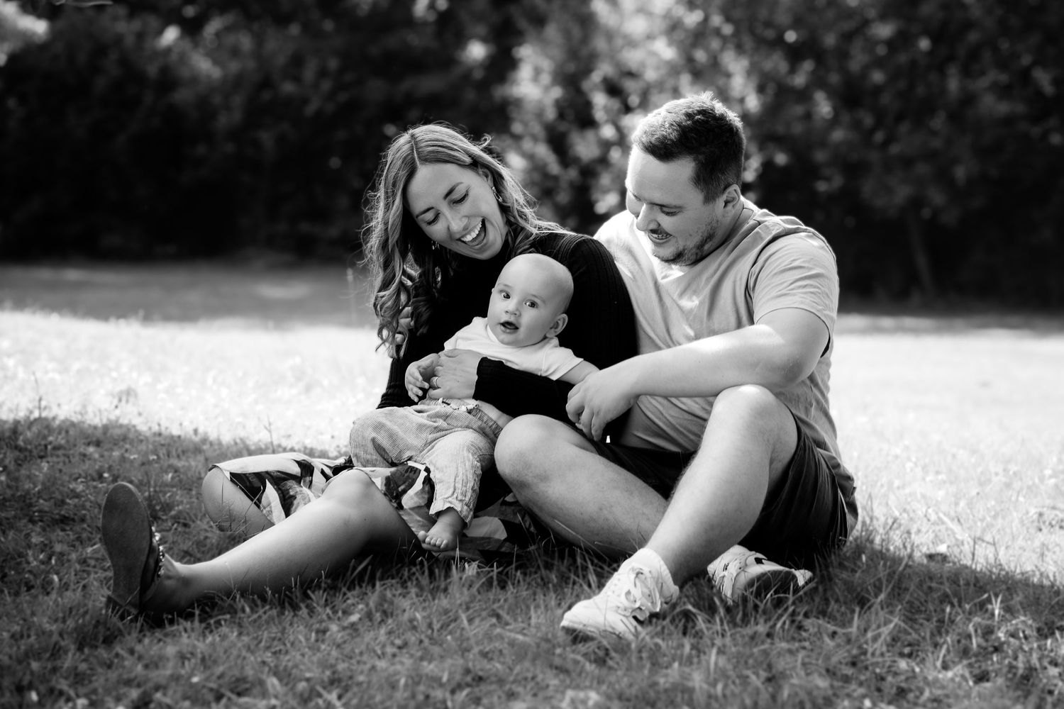 Black and white photo of a happy young family with a baby sitting on grass in a park.