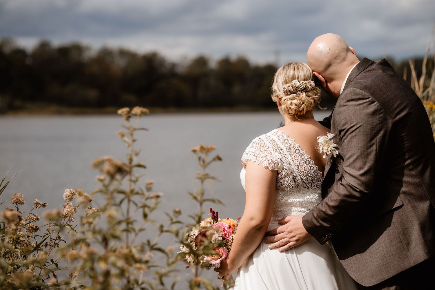 A bride and groom share a romantic moment by a lakeside, with the bride holding a pink bouquet on their wedding day.