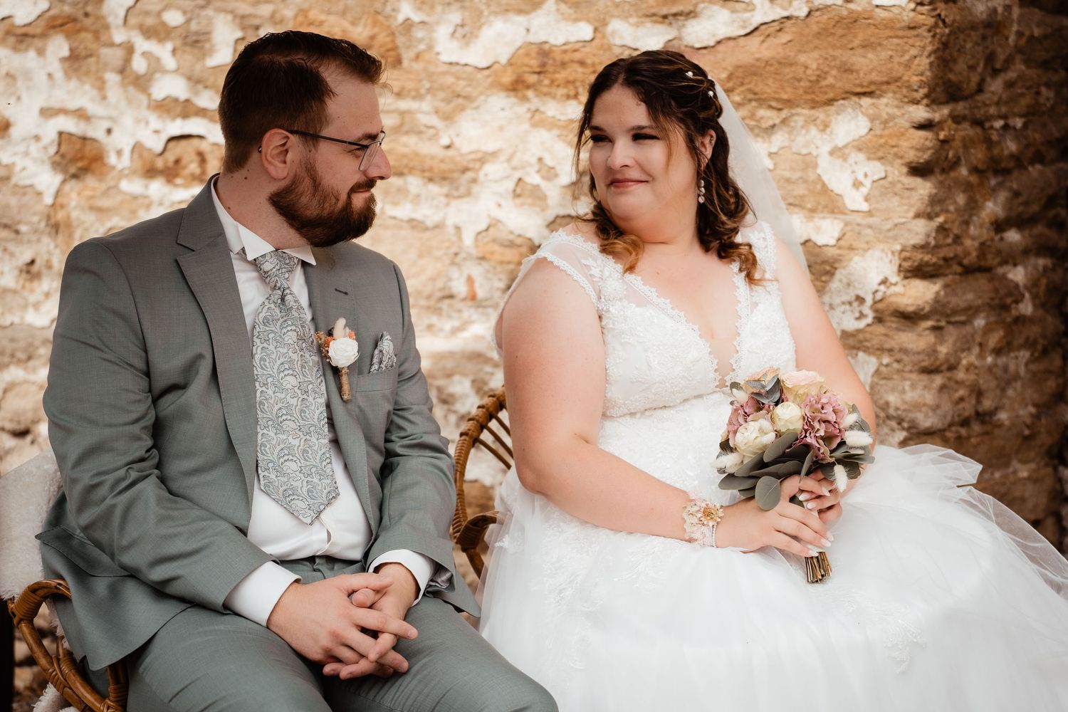 Bride in white gown holding bouquet sits beside groom in gray suit, smiling at each other in rustic stone venue.
