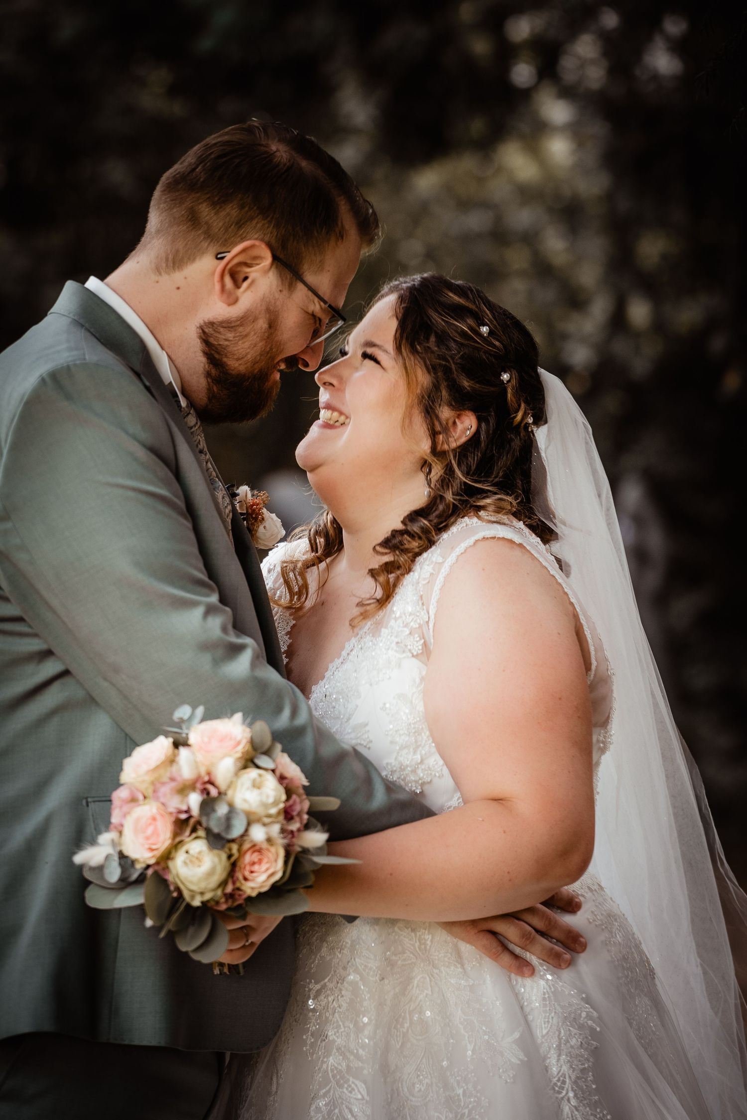 Bride and groom share an intimate moment, smiling at each other, bride holding a pastel bouquet in a white lace gown.