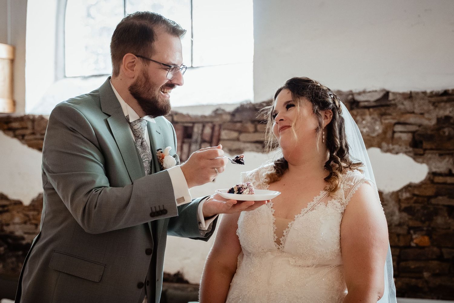 Smiling groom in sage suit feeds wedding cake to bride in lace veil inside rustic stone venue.