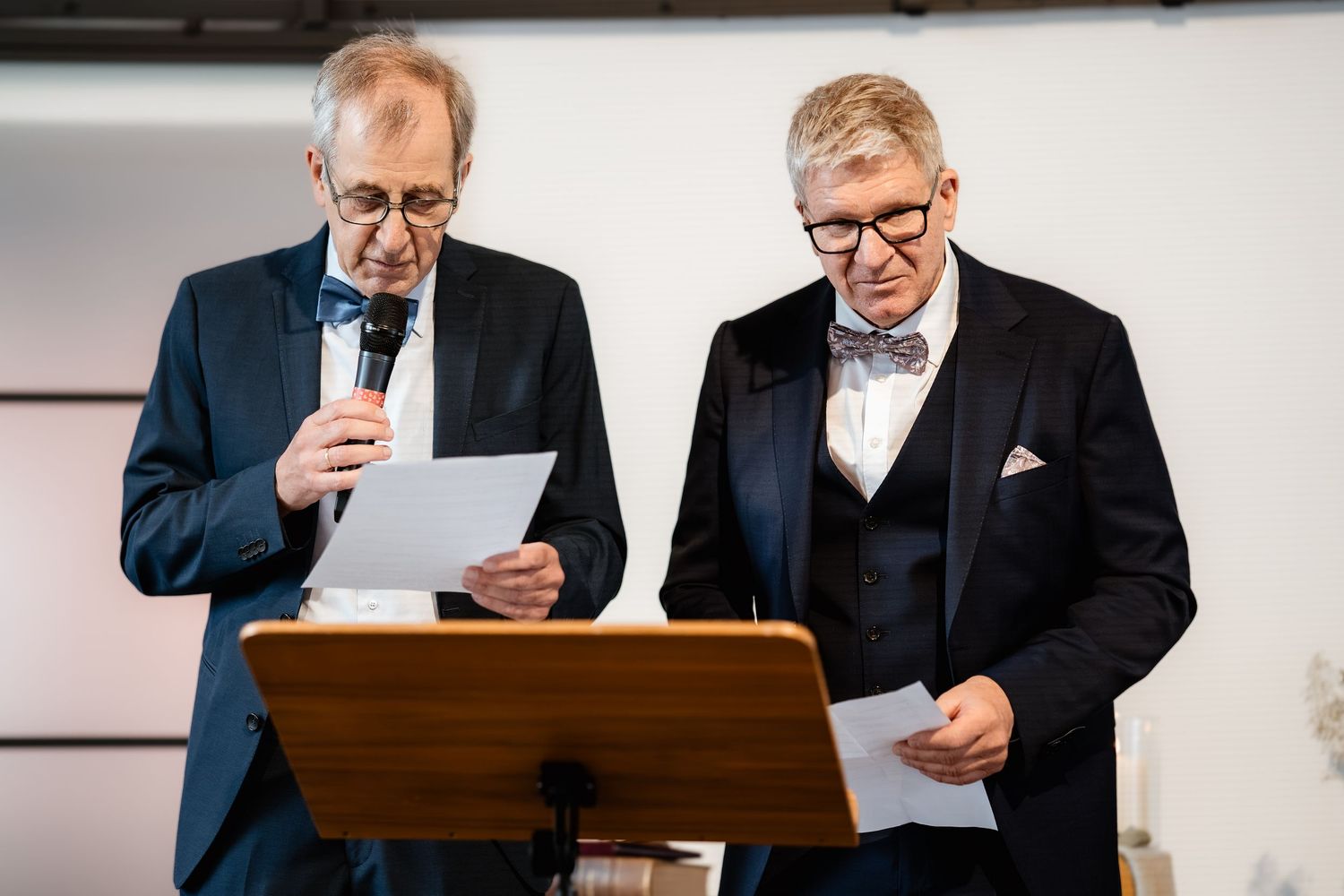 Two elderly men in formal suits read from papers at a podium during a wedding ceremony.