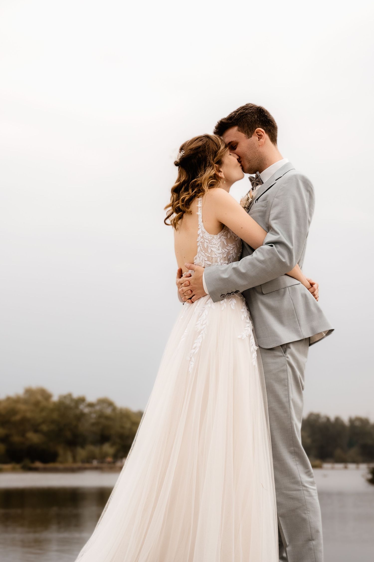 Bride and groom share romantic embrace near lakeside in soft natural light.