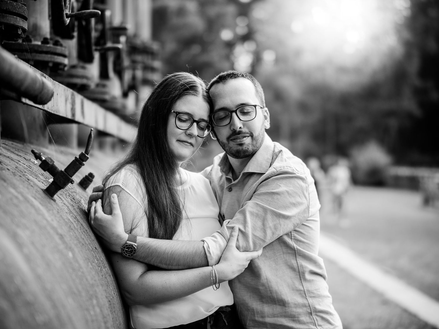 Black and white photo of a couple embracing tenderly near a stone wall, woman with glasses leaning into mans arms.
