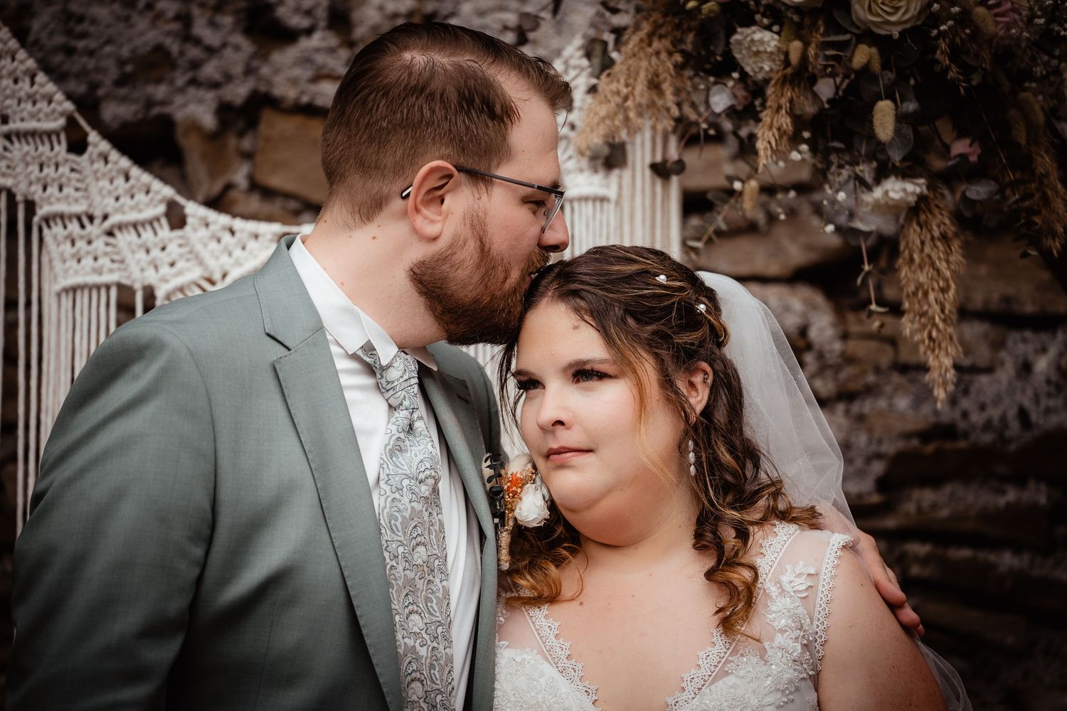Groom tenderly kissing bride on the cheek before a bohemian macrame wedding backdrop.