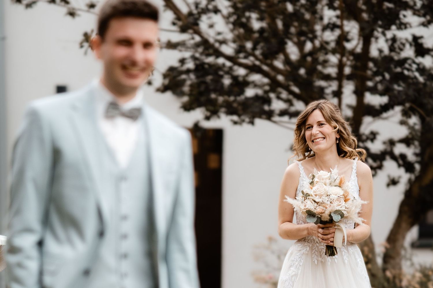 Smiling bride holding bouquet stands behind groom in light blue suit at outdoor wedding ceremony.