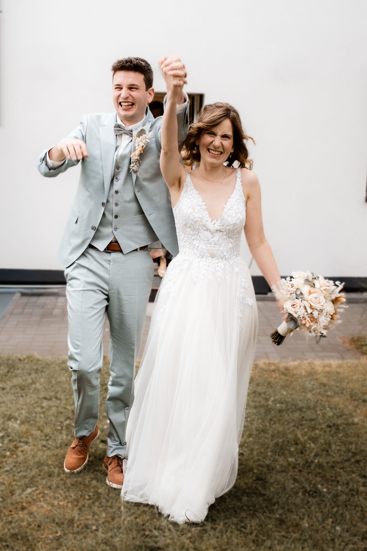 Excited bride and groom celebrate outdoors raising hands with bouquet after wedding ceremony.