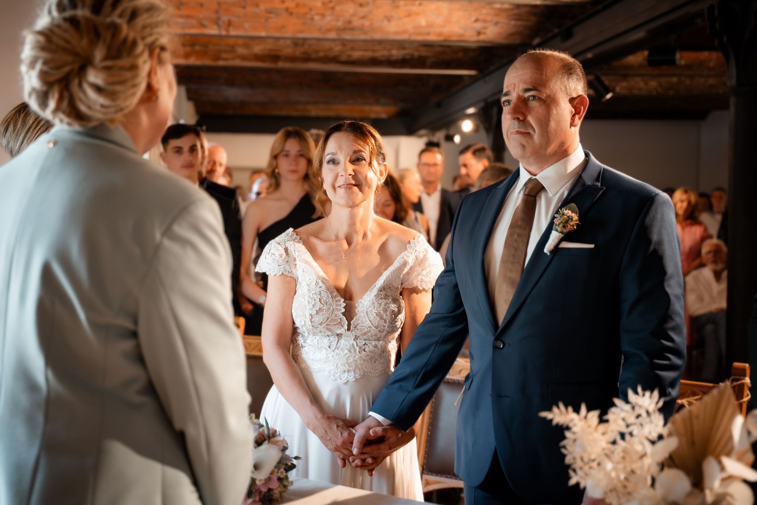 Bride and groom hold hands during wedding ceremony indoors, officiant faces them, guests watch in background.