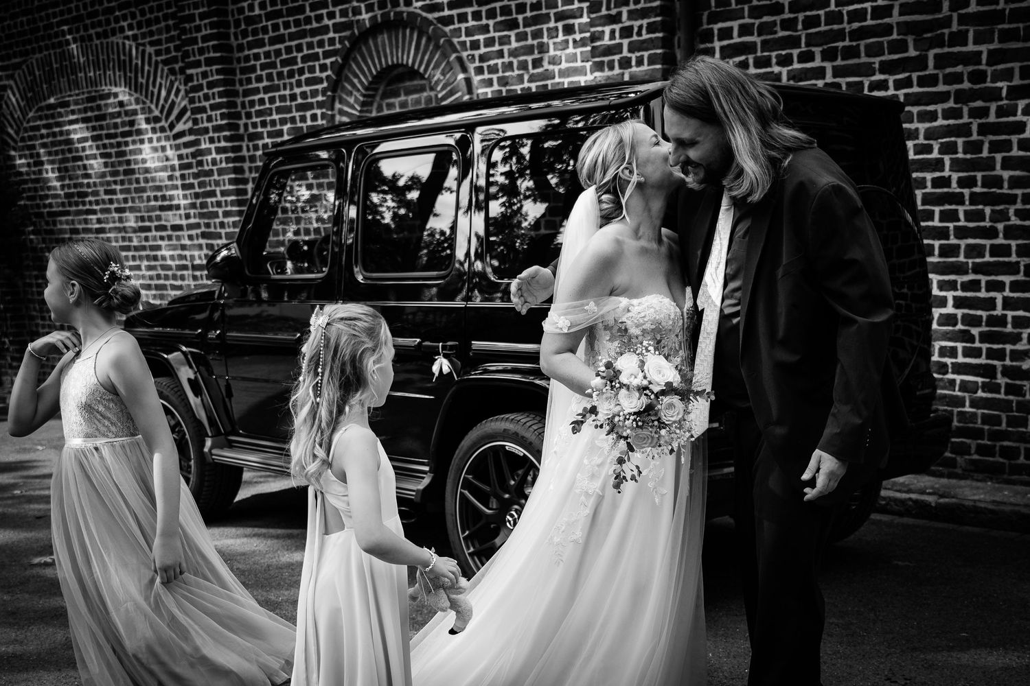 Black and white wedding photo of bride and groom with flower girls near vintage car by brick wall.