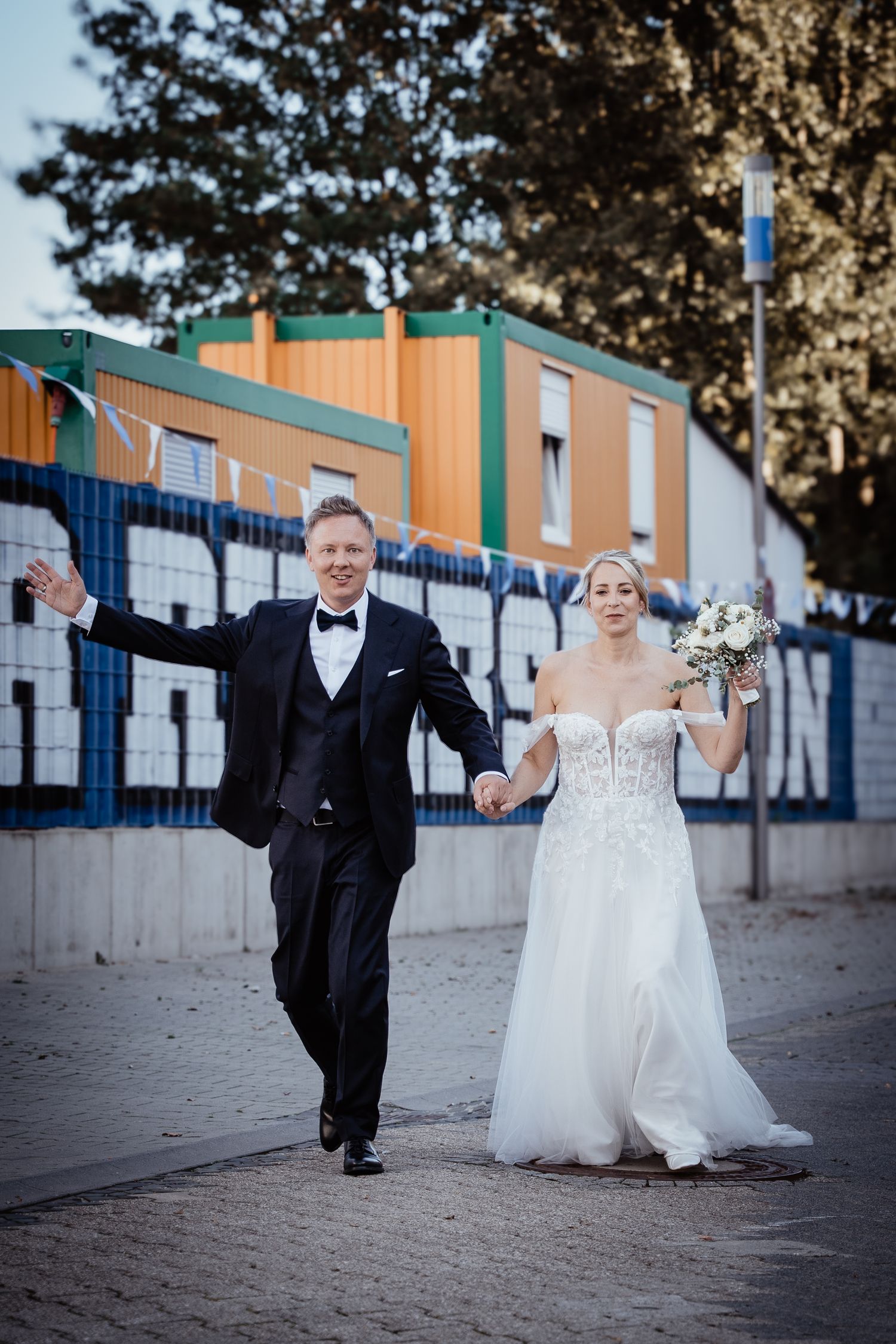 Joyful bride and groom walking hand in hand along a colorful urban street after wedding.