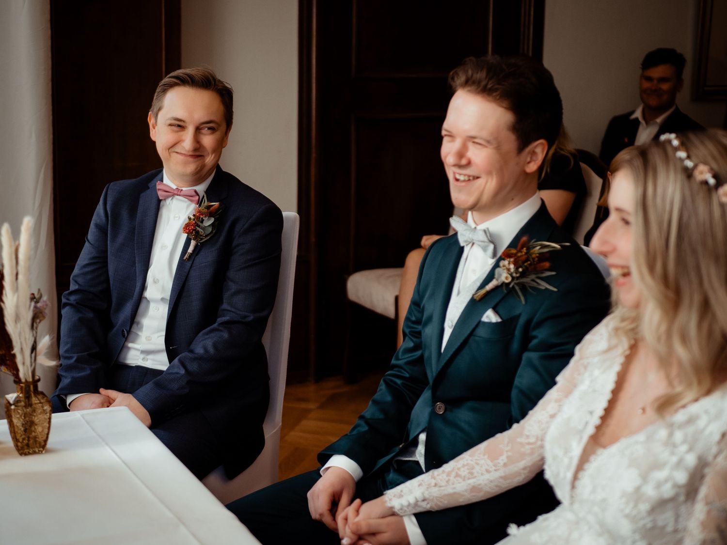 Two grooms smiling during wedding ceremony, one in navy suit, one in green suit, with bride in white dress.