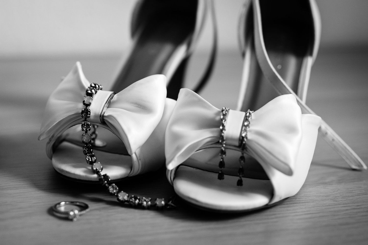 Black and white photo of elegant bridal heels with bow detail, adorned with crystal jewelry and a ring on wooden surface.