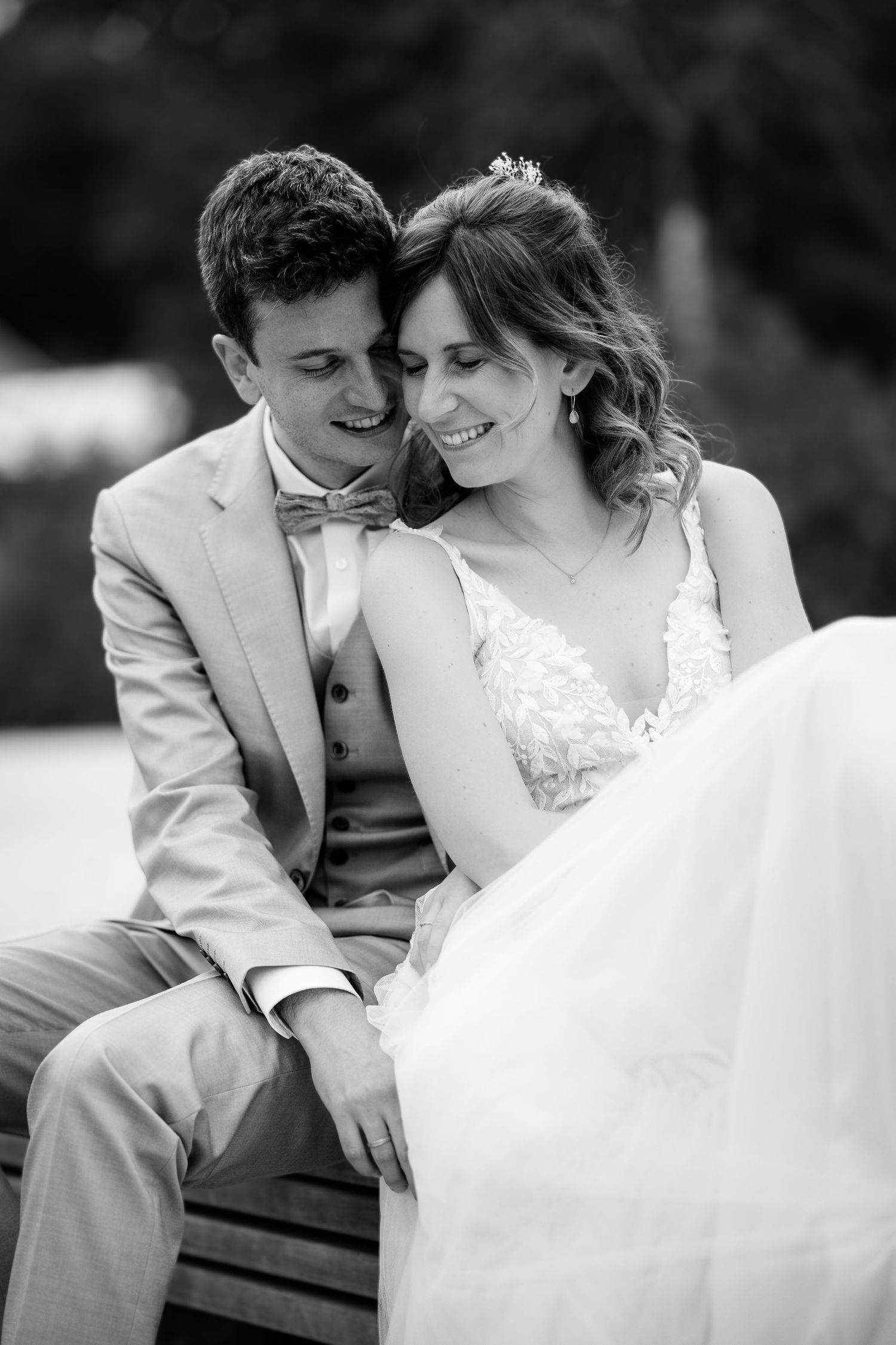 Black and white photo of a smiling bride and groom sitting closely together on a bench in an outdoor setting.