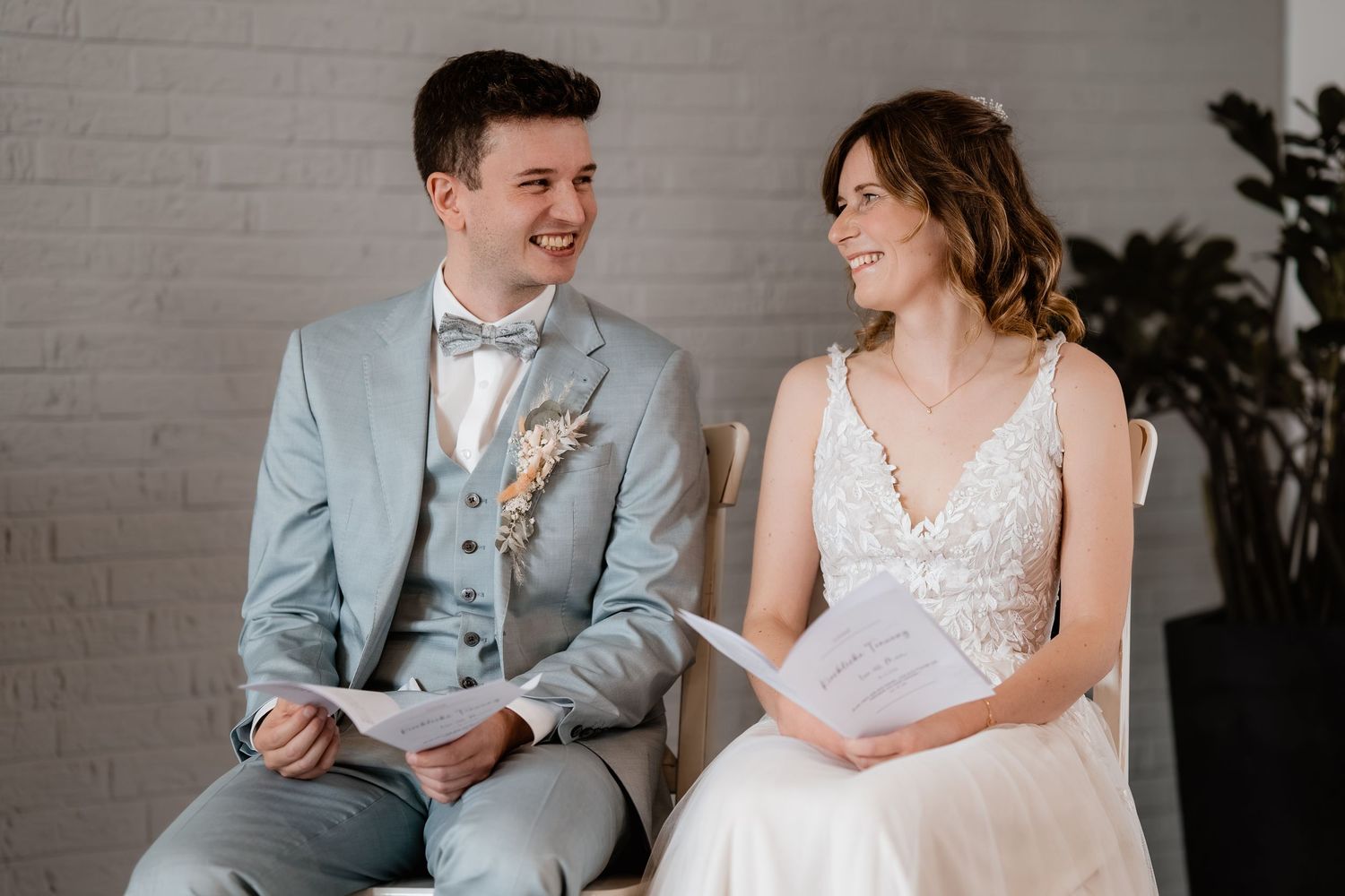 Smiling bride and groom in gray suit exchanging vows while seated, holding papers at wedding ceremony.