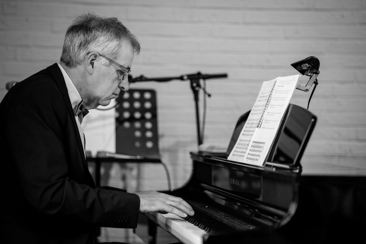 Elderly man playing grand piano in black and white, reading sheet music at an intimate indoor performance.