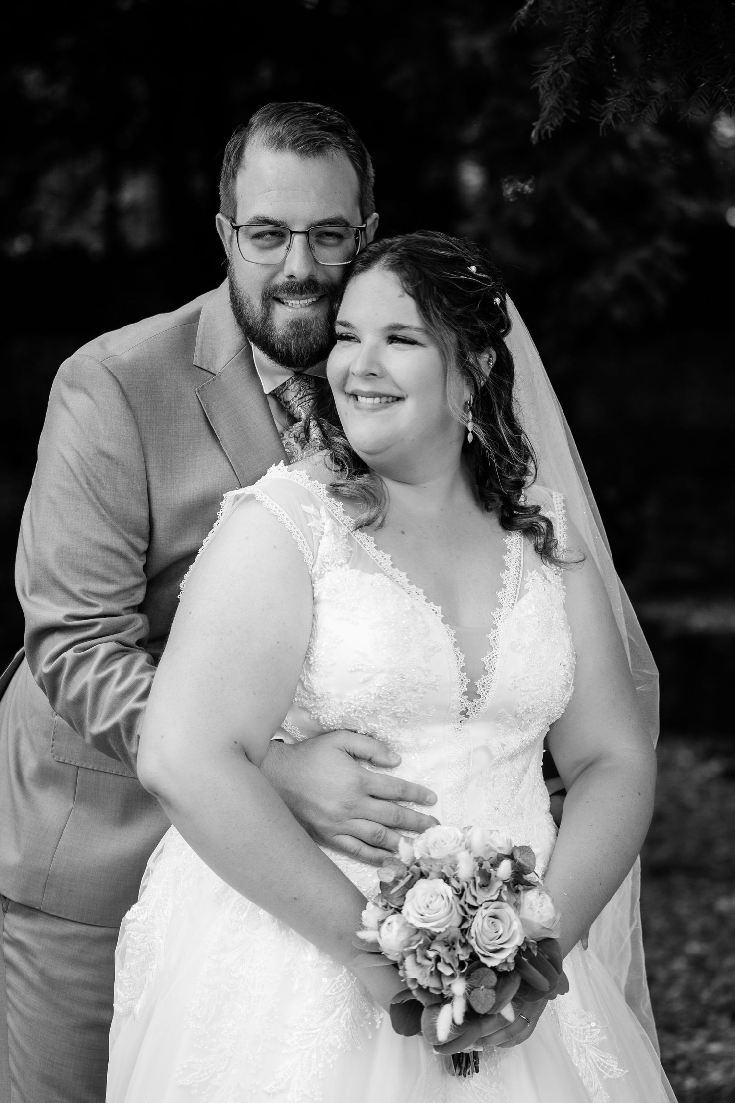 Black and white wedding portrait of smiling bride holding bouquet with groom embracing her from behind.