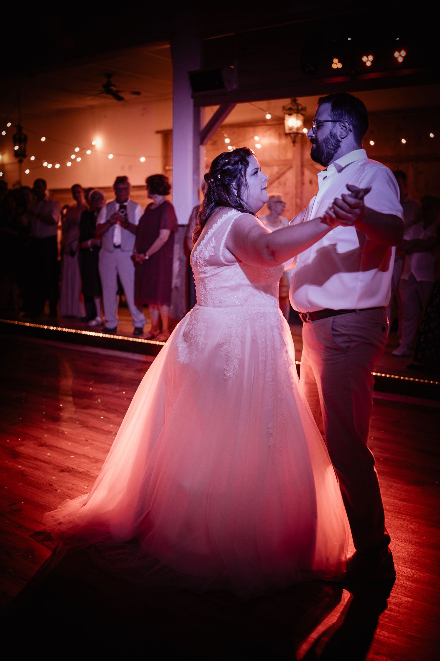 Bride and groom sharing their first dance under warm pink lights with guests watching.