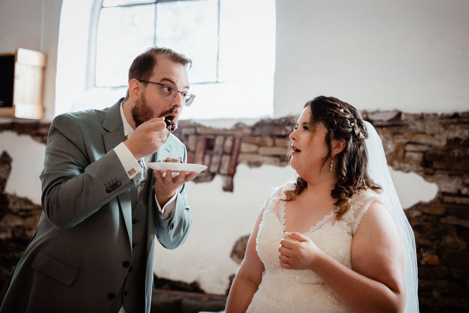 Groom in grey suit feeds wedding cake to surprised bride in lace dress at rustic indoor reception.