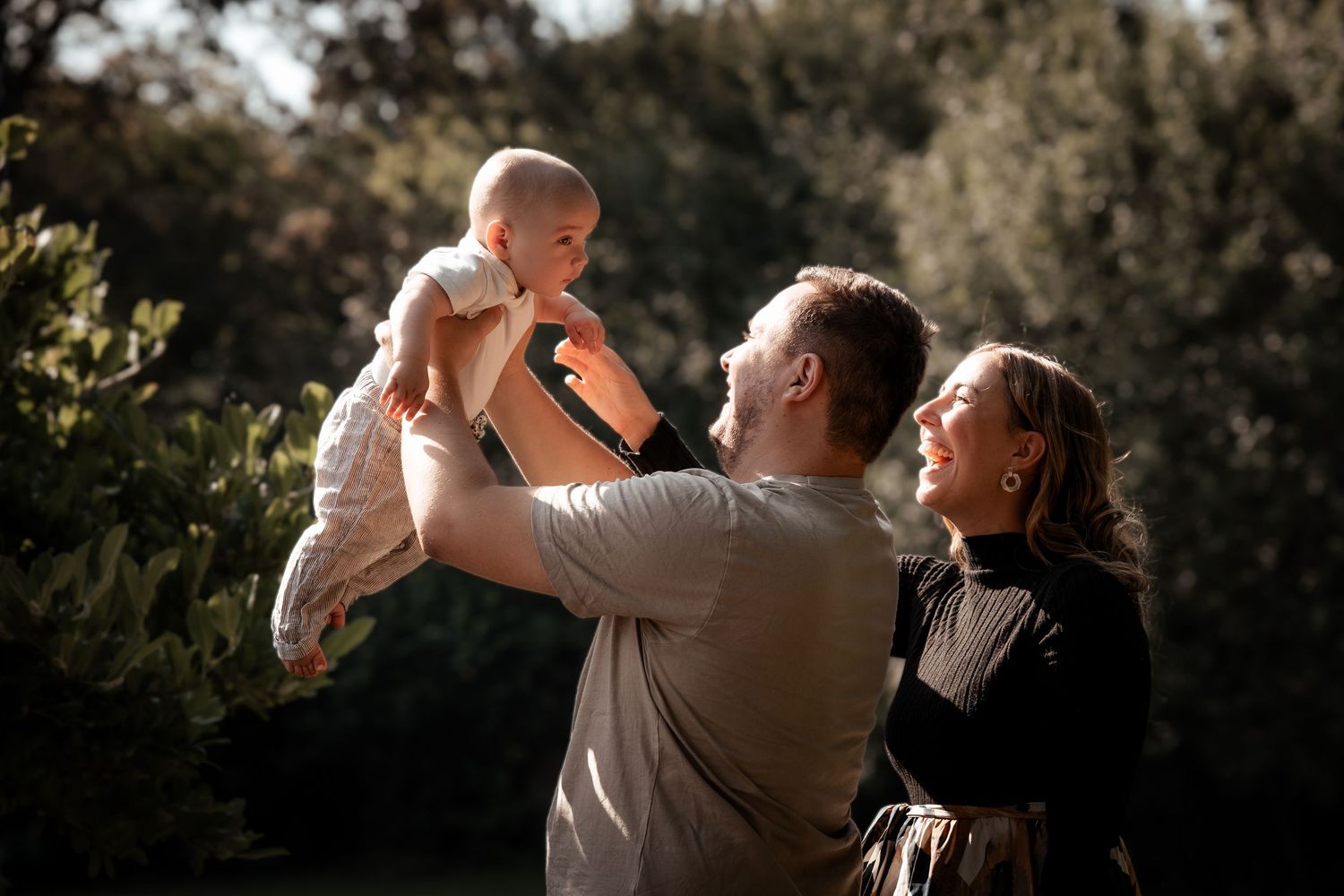 Happy family outdoors, father lifting smiling baby in air while mother watches lovingly in sunny garden.
