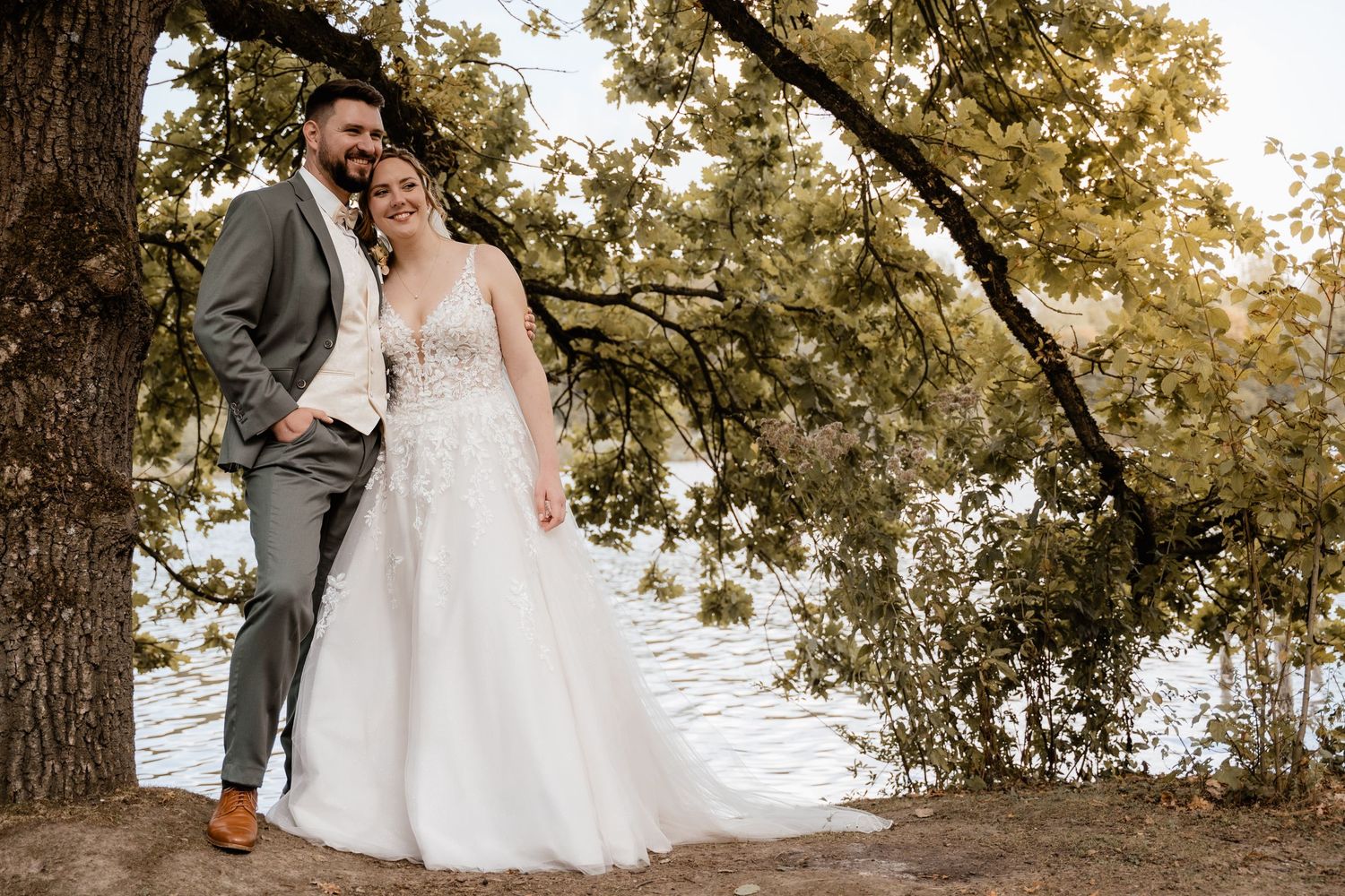 Bride and groom pose romantically under a tree by the water in elegant wedding attire.