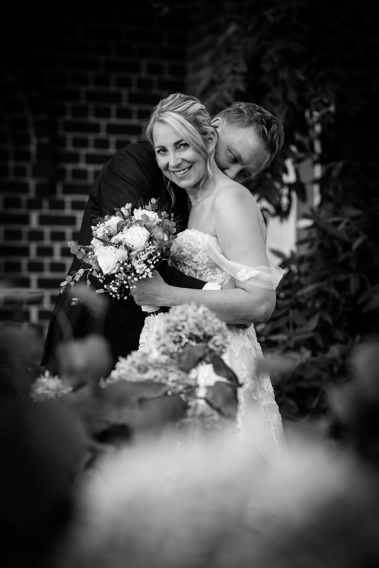 Black and white photo of groom embracing bride holding bouquet in a garden setting.