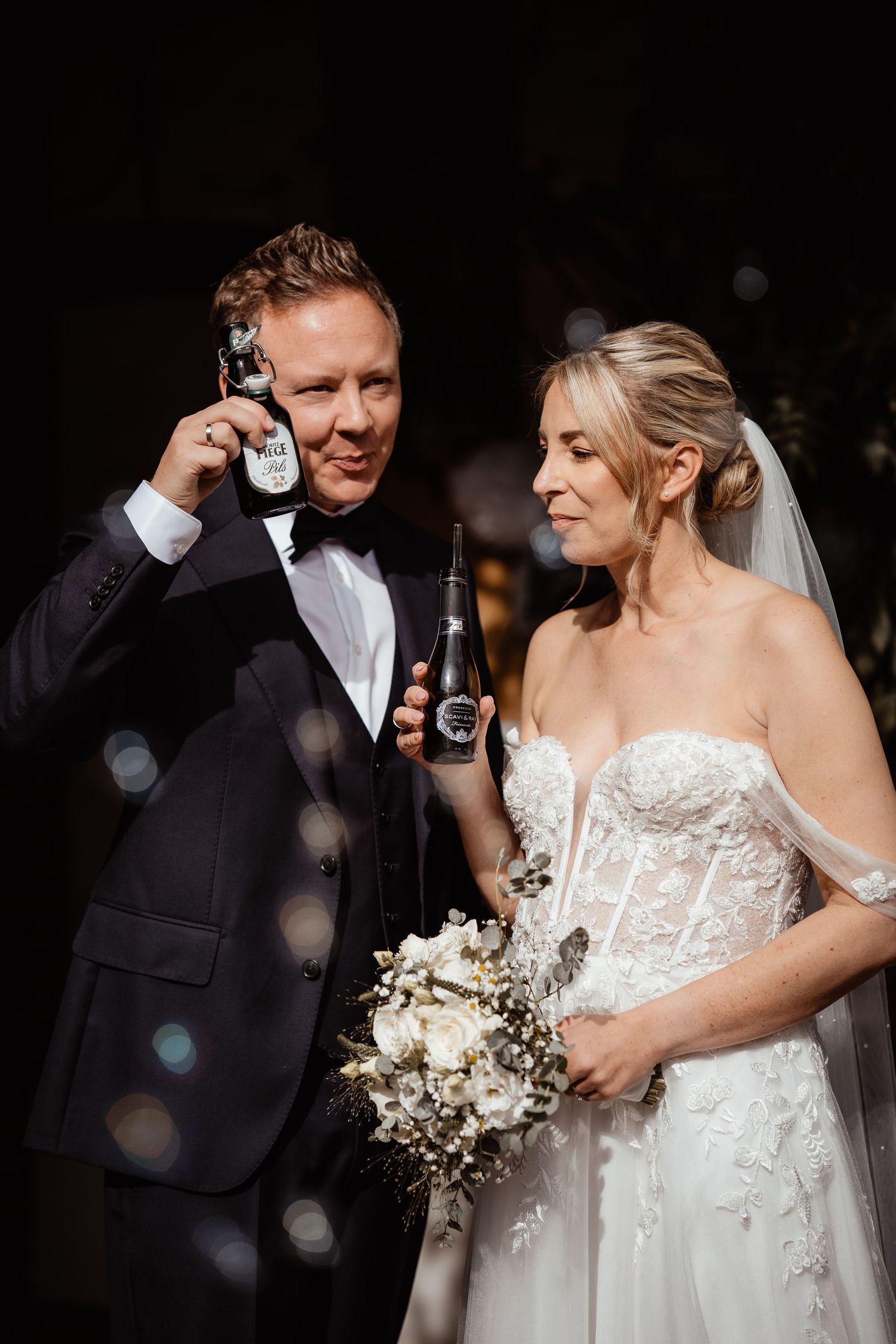 Smiling groom in tuxedo holds beer bottle to ear while bride holds bouquet at wedding.