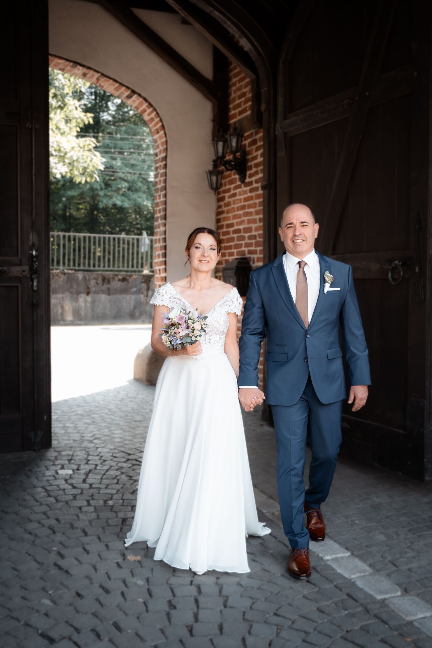 Smiling bride and groom walking hand in hand through historic brick archway on wedding day.