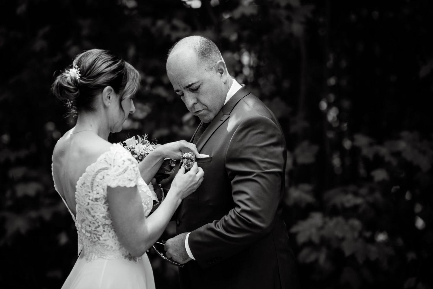 Bride pinning boutonniere on groom in black and white wedding photo among lush greenery.