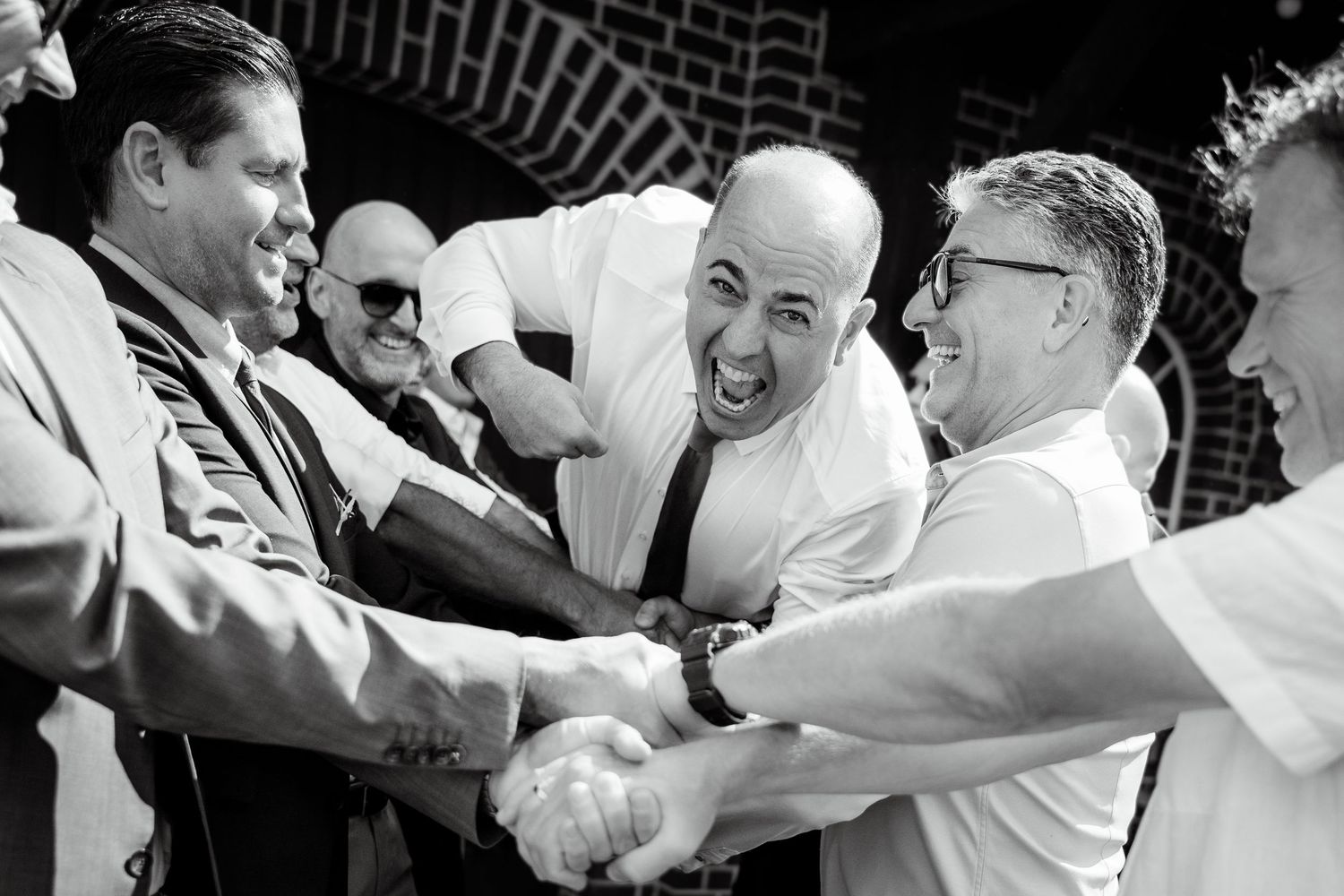 Group of men laughing and shaking hands in lively black and white candid wedding celebration photo.