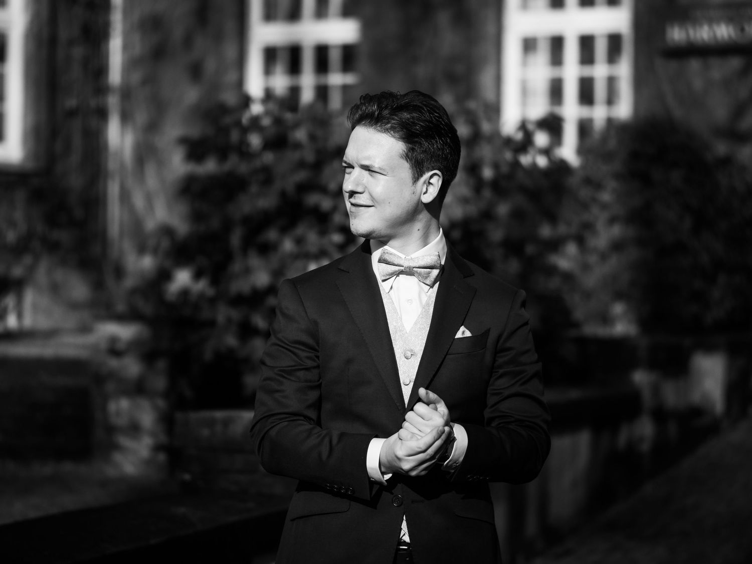 Elegant man in black tuxedo and bow tie poses outdoors in black and white portrait photo.